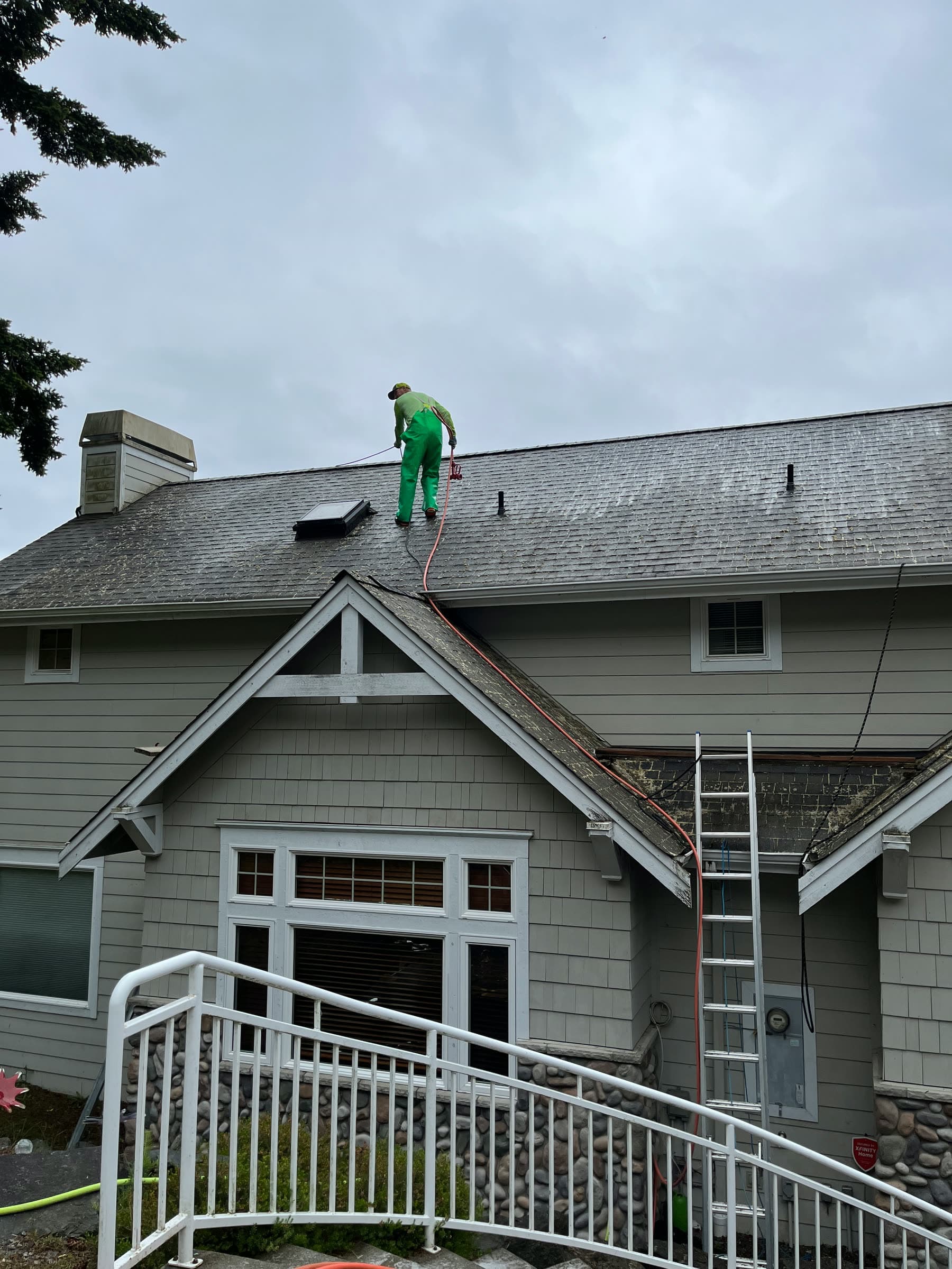A crew member in green on a grey shingle roof, working a hose line down from the ridge, a ladder against the house below.