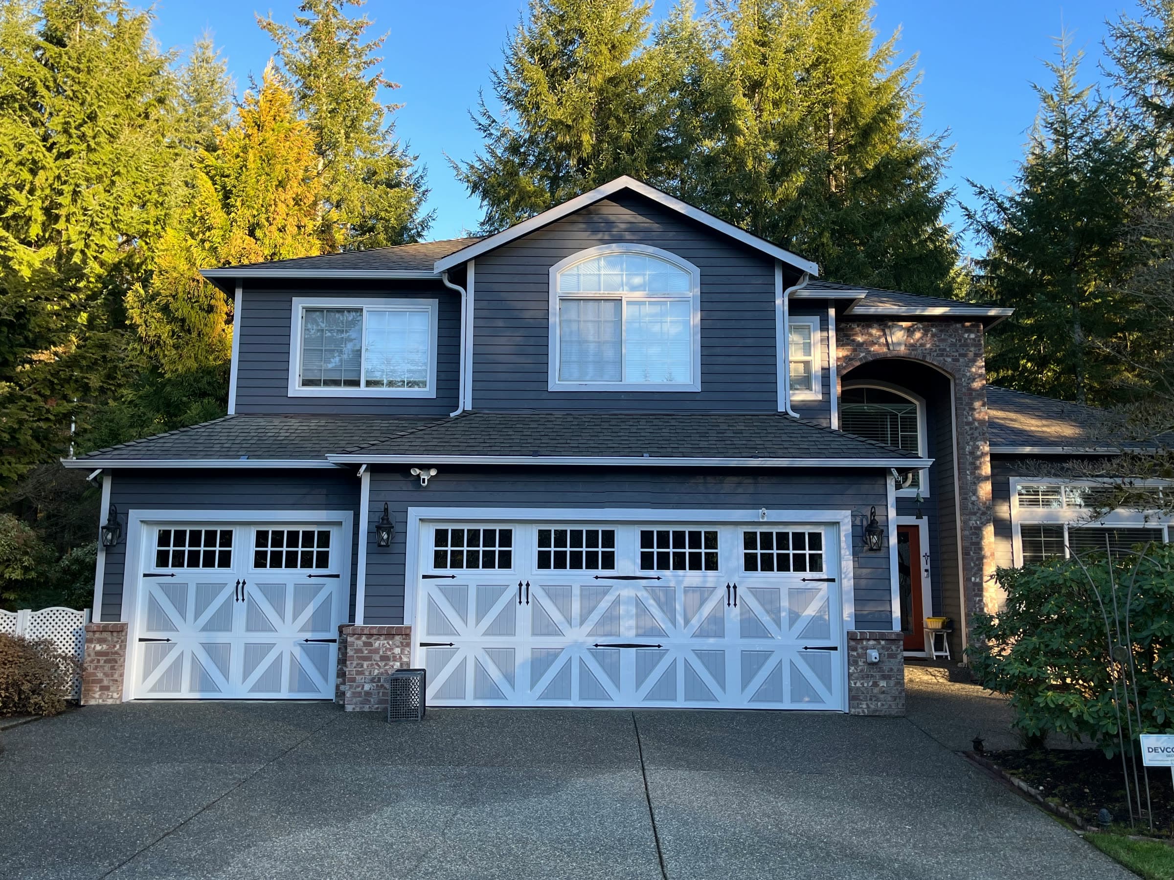 A blue two-storey house with white carriage garage doors in golden evening light, tall firs behind it.