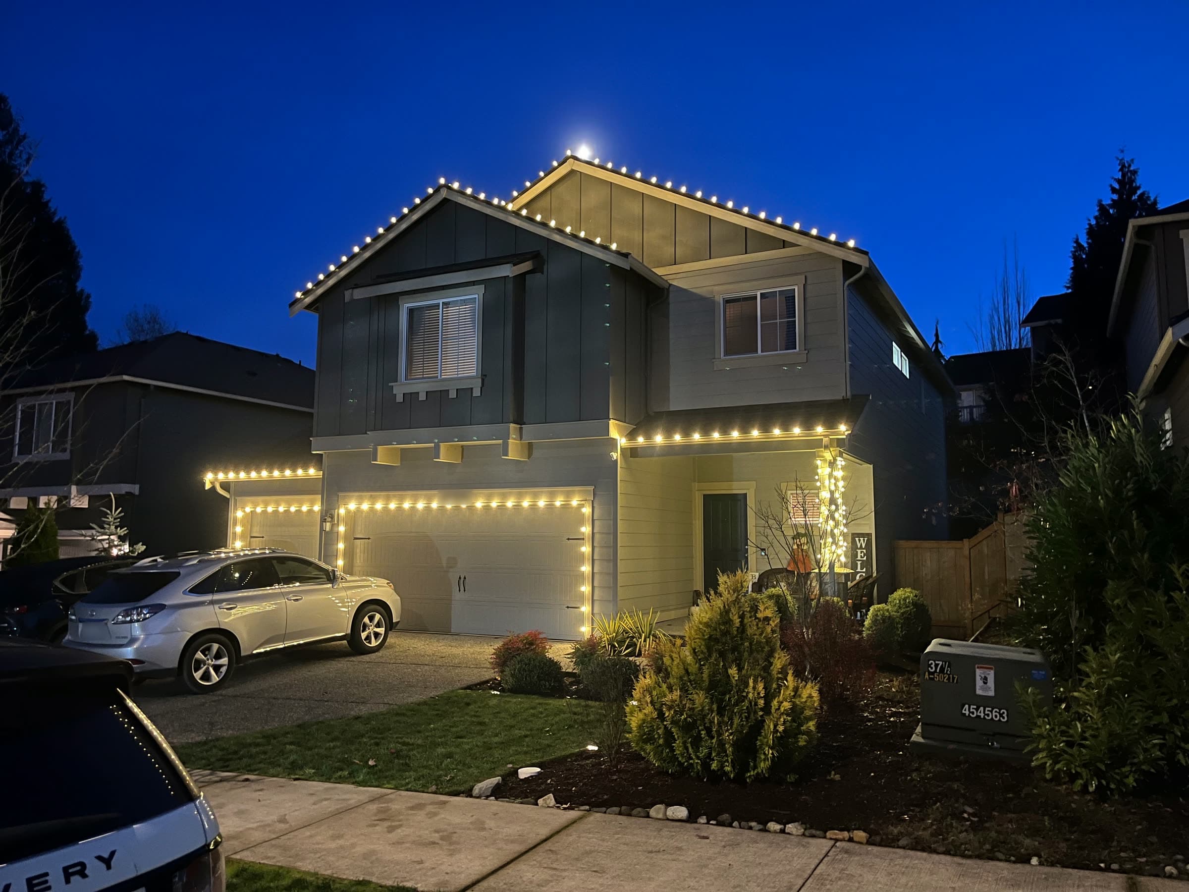 A two-storey house outlined in warm white Christmas lights at blue hour.