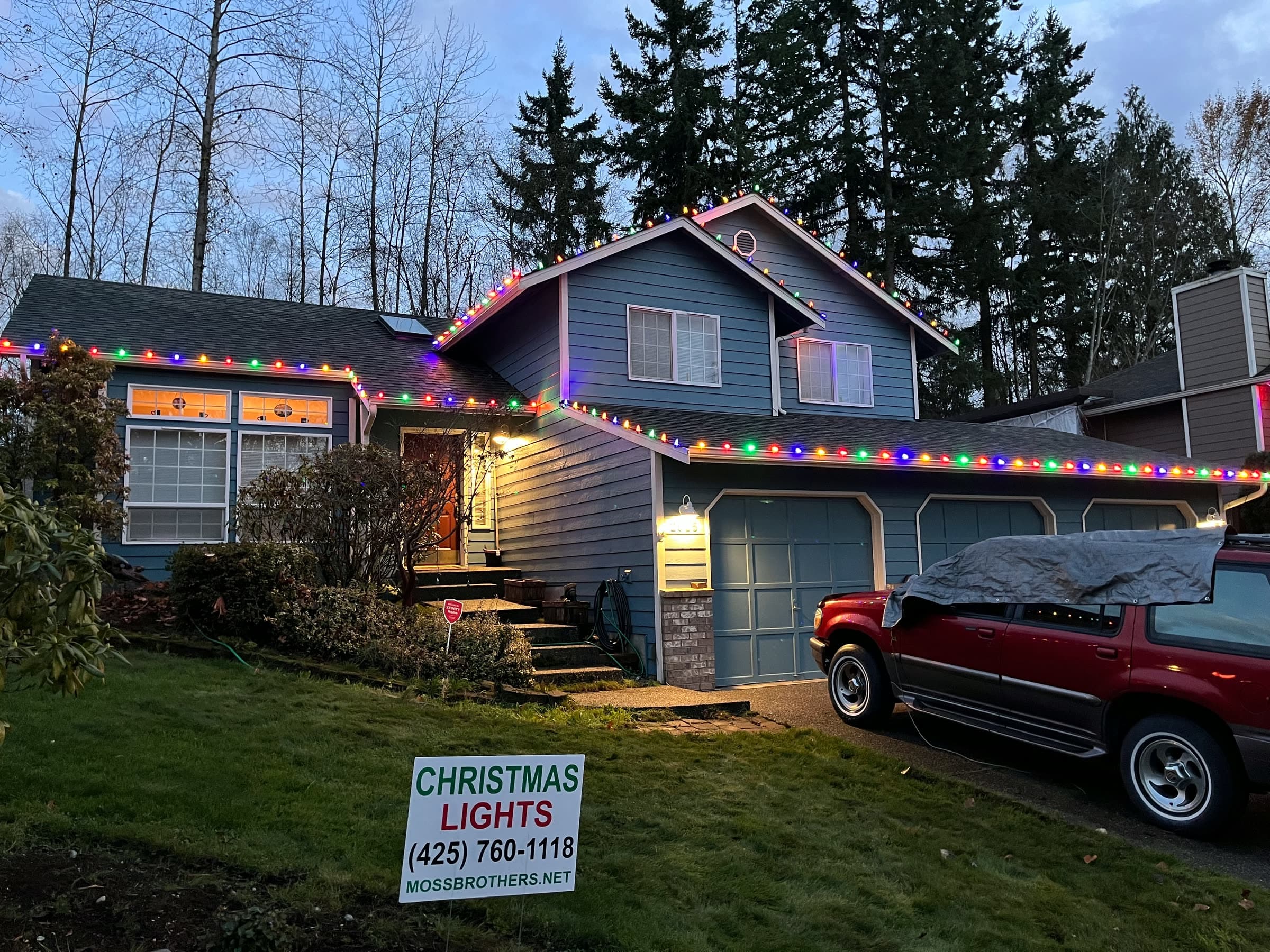 A blue house with multicolor lights along the roofline and a Mossbrothers Christmas lights sign in the yard.