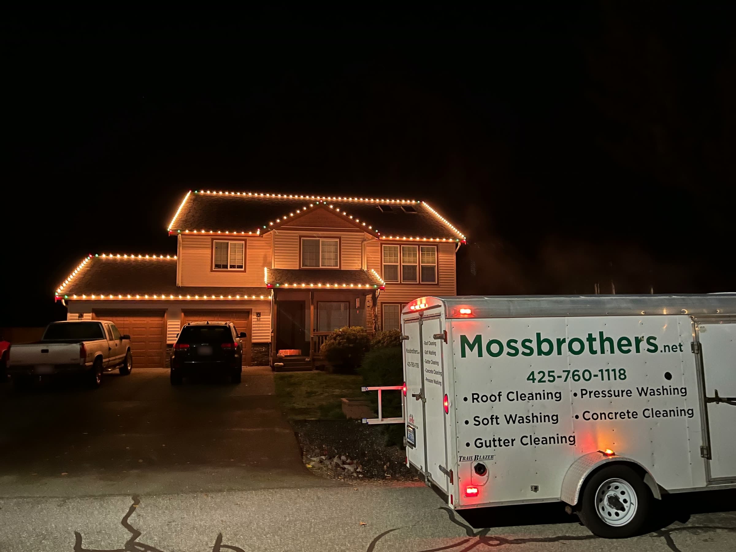 A house lit with warm white Christmas lights at night, the Mossbrothers trailer parked in front.
